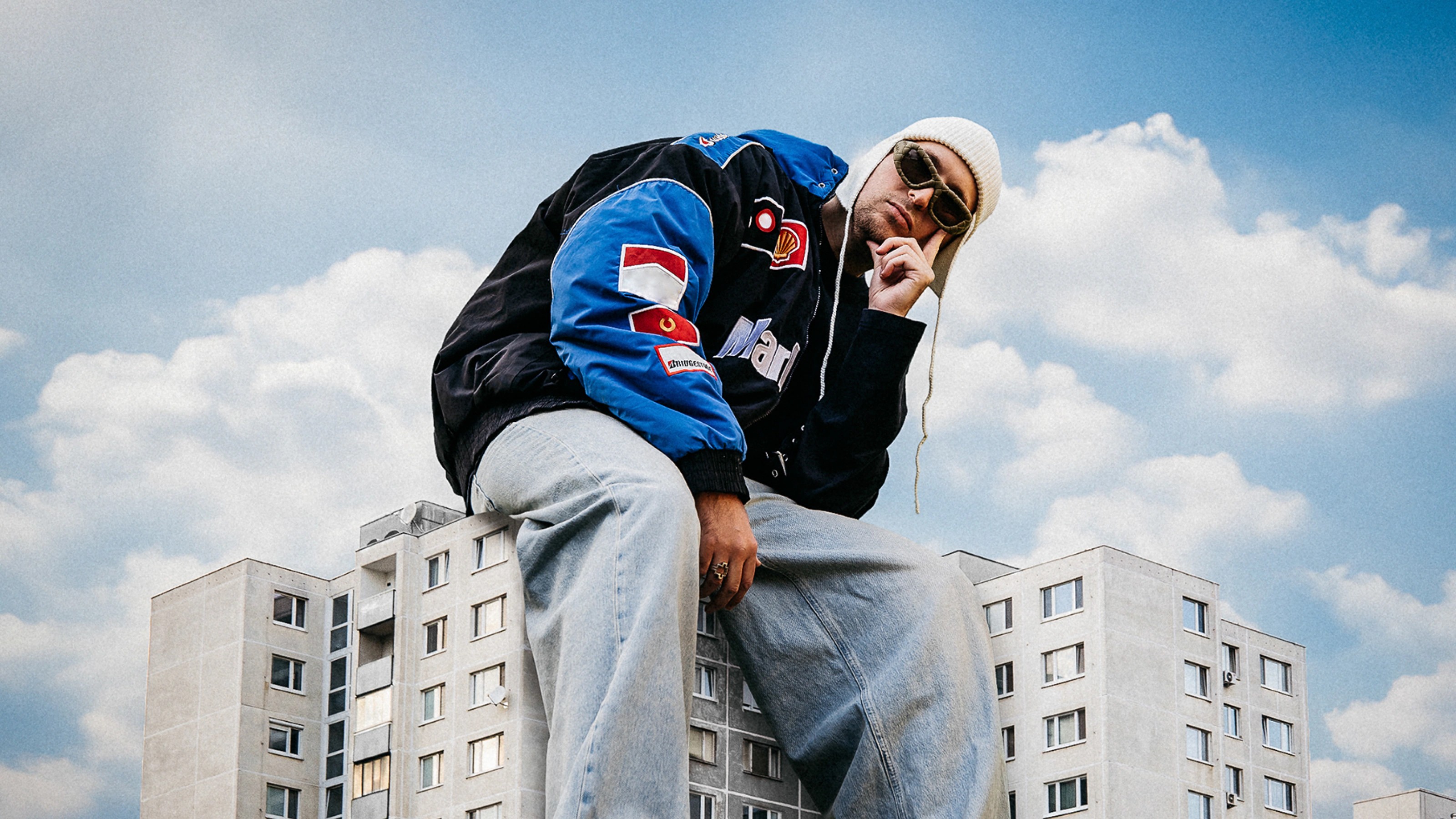 Gleb sitting on the edge of a rooftop above panel-block apartment buildings, blue sky behind him
